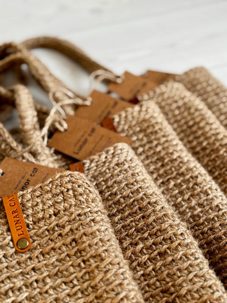 Puede incluir: Primer plano de varias bolsas de mano de yute tejido. Cada bolsa tiene un asa a juego y una etiqueta marr&oacute;n con el texto "LUNAR CAT" impreso. Las bolsas son de color marr&oacute;n natural y parecen hechas a mano.