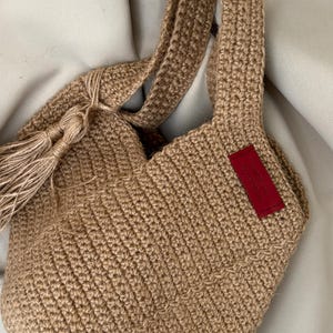 May include: A hand-crocheted tote bag in a natural tan color. The bag features sturdy handles and a decorative tassel. A small red tag is sewn on the side. The bag is made of a thick, textured yarn, perfect for carrying items.