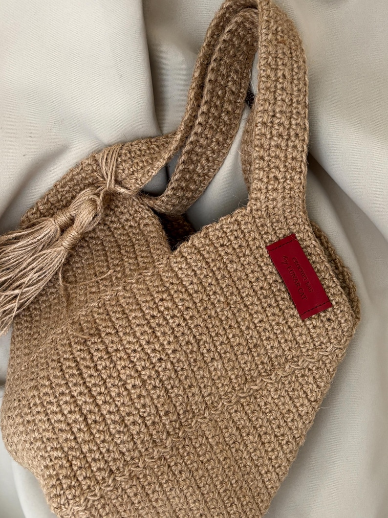May include: A hand-crocheted tote bag in a natural tan color. The bag features sturdy handles and a decorative tassel. A small red tag is sewn on the side. The bag is made of a thick, textured yarn, perfect for carrying items.