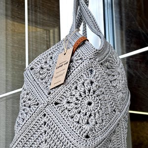 May include: A gray crochet bag featuring a square motif pattern with a central floral design. The bag has a twisted handle and a brown leather-like tag attached with twine, displaying the text "CROCHET BAG by Lunar cat" in black ink.