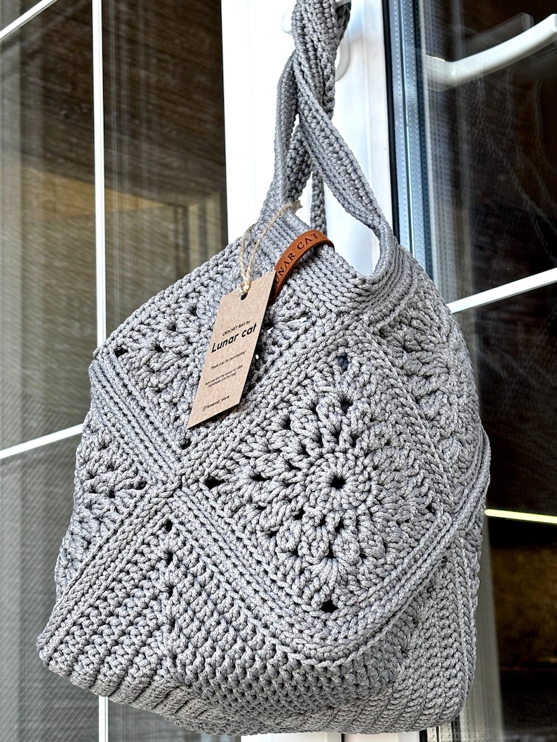 May include: A gray crochet bag featuring a square motif pattern with a central floral design. The bag has a twisted handle and a brown leather-like tag attached with twine, displaying the text "CROCHET BAG by Lunar cat" in black ink.