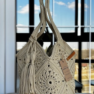 May include: A beige crochet tote bag featuring a square motif pattern, two long shoulder straps, and a decorative tassel accent. A brown leather-like tag is attached to the side, and a rectangular paper hang tag displays the text Lunar cat.
