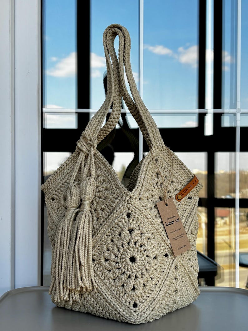 May include: A beige crochet tote bag featuring a square motif pattern, two long shoulder straps, and a decorative tassel accent. A brown leather-like tag is attached to the side, and a rectangular paper hang tag displays the text Lunar cat.