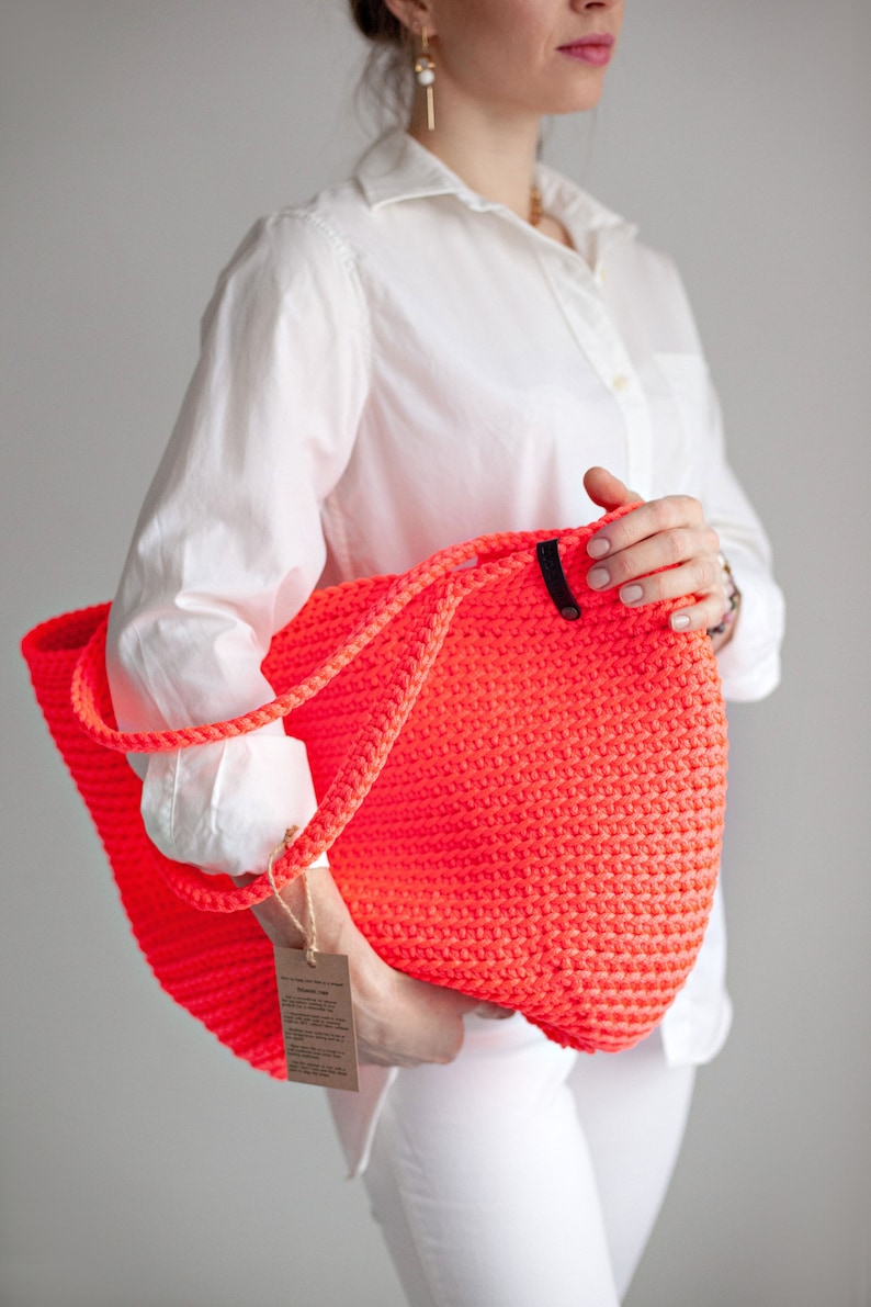 May include: A bright neon pink crocheted tote bag with black leather handles. The bag is large and roomy, perfect for carrying all your essentials.