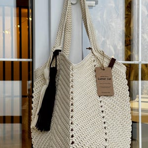 May include: Cream-colored crochet tote bag with a black tassel and a tag that reads "Lunar cat". The bag has a chevron pattern and long straps. The bag is hanging on a white door handle.