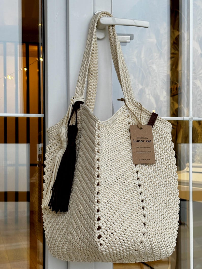 May include: Cream-colored crochet tote bag with a black tassel and a tag that reads "Lunar cat". The bag has a chevron pattern and long straps. The bag is hanging on a white door handle.