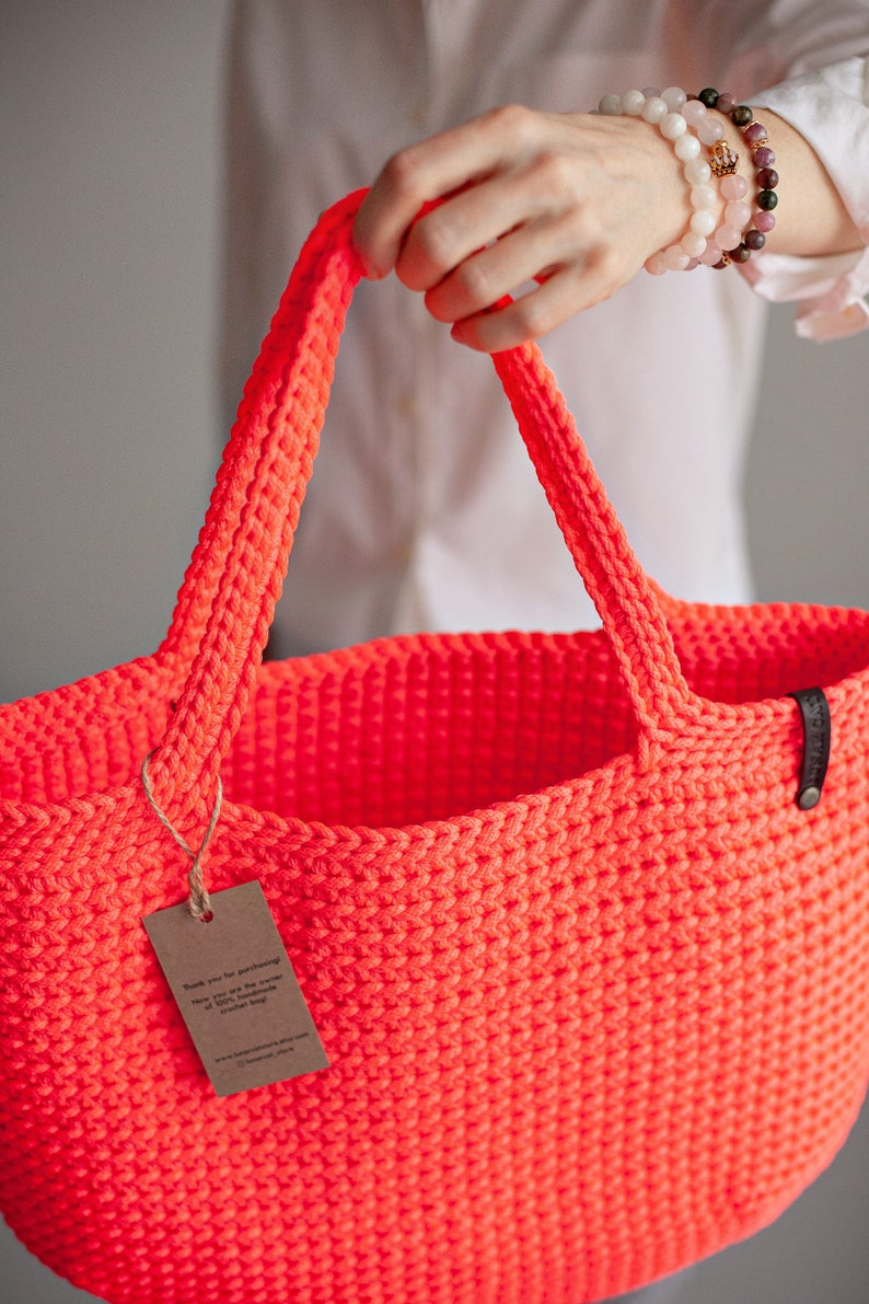 May include: A bright neon orange crocheted tote bag with a brown tag that says "Thank you for supporting the power of 100% handmade crochet bags".