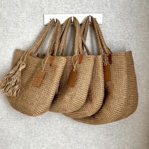 Puede incluir: Cuatro bolsas de mano de yute tejido con asas resistentes cuelgan de un soporte blanco contra una pared gris. Cada bolsa tiene forma rectangular, una etiqueta de cuero marr&oacute;n y una borla de yute a juego. Las bolsas son de color natural.