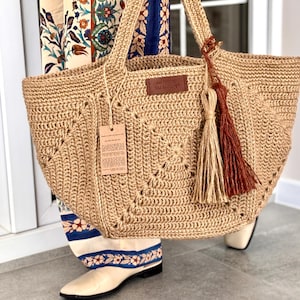 Peut inclure: Un grand sac fourre-tout beige crocheté avec des anses robustes et un pompon décoratif. Le sac présente un motif carré et une petite étiquette en cuir. Une étiquette de prix est suspendue à la poignée. Le sac est porté par quelqu'un portant des vêtements à motifs et des chaussures crème.