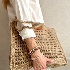 May include: A large, brown, crocheted tote bag with a leather handle. The bag is made of natural fibres and has a square shape. The bag is being held by a person wearing a white shirt and white trousers.