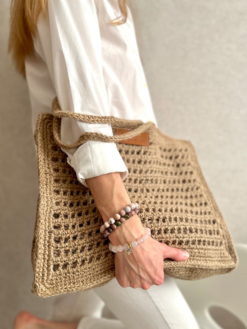 May include: A large, brown, crocheted tote bag with a leather handle. The bag is made of natural fibres and has a square shape. The bag is being held by a person wearing a white shirt and white trousers.