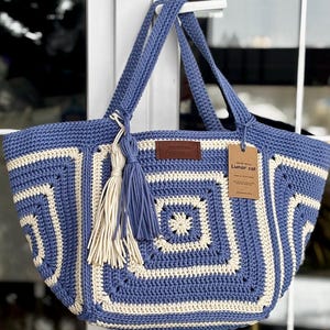 Puede incluir: Un bolso tote de crochet azul y crema con un diseño de patrón cuadrado. El bolso tiene dos asas y una borla. Una etiqueta de cuero marrón y una etiqueta blanca con el texto "Lunar cat" están adheridas al bolso.
