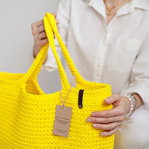 May include: A bright yellow crocheted tote bag with a brown leather handle and a small brown tag attached. The bag is being held by a person wearing a white shirt.
