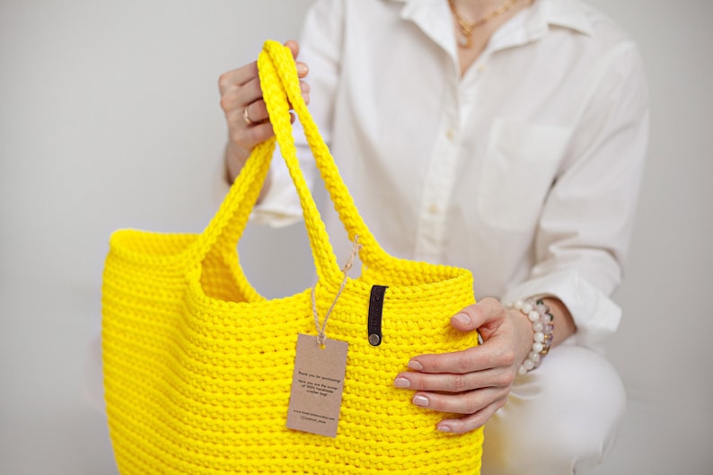 May include: A bright yellow crocheted tote bag with a brown leather handle and a small brown tag attached. The bag is being held by a person wearing a white shirt.