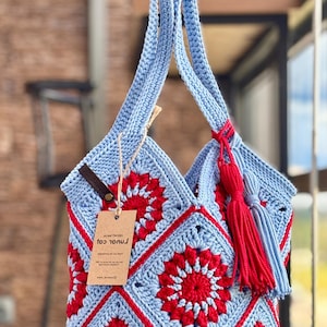 May include: A light blue and red crochet tote bag constructed from square motifs featuring a central red floral pattern. The bag includes a long shoulder strap, a red and light blue tassel accessory, and a brown tag with printed text attached.