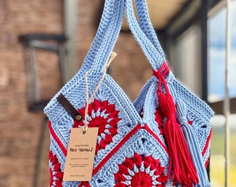 May include: A light blue and red crochet tote bag constructed from square motifs featuring a central red floral pattern. The bag includes a long shoulder strap, a red and light blue tassel accessory, and a brown tag with printed text attached.