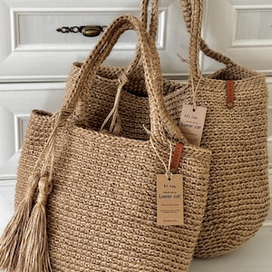 Puede incluir: Dos bolsas de mano de yute tejido en color beige natural, con asas resistentes y borlas. Las bolsas tienen etiquetas que dicen "Lunar Cat". Se exhiben frente a un armario blanco.