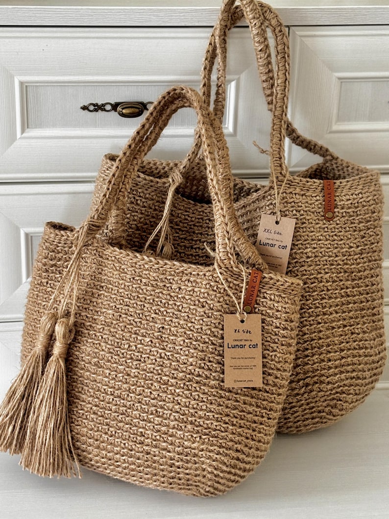 Puede incluir: Dos bolsas de mano de yute tejido en color beige natural, con asas resistentes y borlas. Las bolsas tienen etiquetas que dicen "Lunar Cat". Se exhiben frente a un armario blanco.
