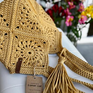 May include: Mustard yellow crochet bag with a square pattern and a long strap. The bag features a tassel and a small leather tag. A paper tag with the text "Lunar cat" is attached to the bag.