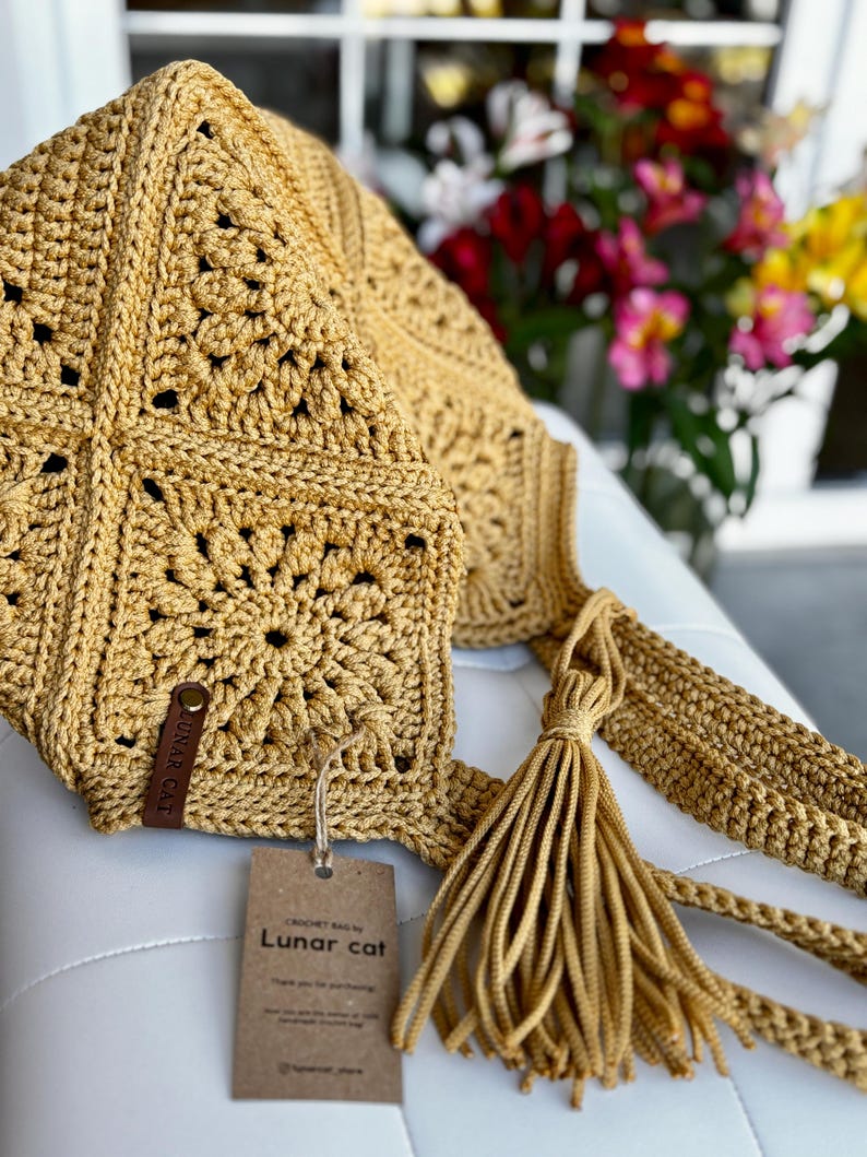 May include: Mustard yellow crochet bag with a square pattern and a long strap. The bag features a tassel and a small leather tag. A paper tag with the text "Lunar cat" is attached to the bag.