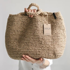 Puede incluir: Un bolso tote grande de yute tejido, de color beige natural, con forma redondeada y un asa resistente. El bolso tiene una etiqueta que dice "Lunar cat". El bolso es sostenido por una persona.