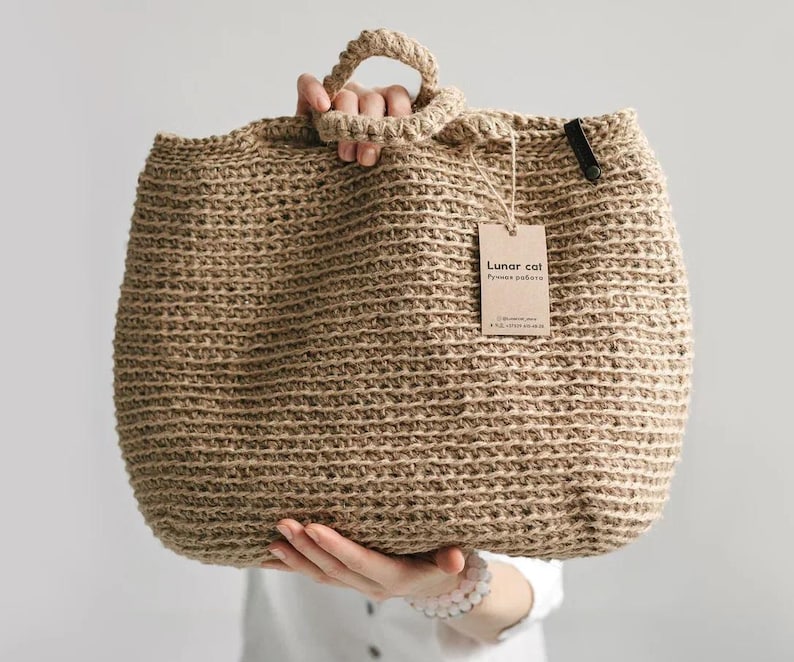 Puede incluir: Un bolso tote grande de yute tejido, de color beige natural, con forma redondeada y un asa resistente. El bolso tiene una etiqueta que dice "Lunar cat". El bolso es sostenido por una persona.