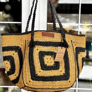 以下が含まれることがあります： 黒と金の配色が印象的な、手作りの大きなかぎ針編みトートバッグ。バッグは幾何学的な正方形のパターンと、黒色の編み込みハンドルが特徴です。「Lunar cat」の文字が入った革製のタグと紙製のタグが付いています。