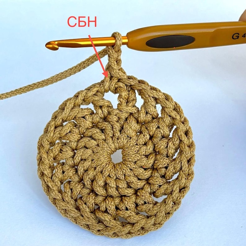 May include: A close-up of a crochet project in progress, featuring a circular design in a warm, tan color. A crochet hook is inserted into the work, with the letters "СБН" visible in red. The image highlights the texture and technique of the craft.