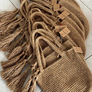 Puede incluir: Un conjunto de bolsas de mano de yute tejido. Cada bolsa tiene un asa trenzada y una borla decorativa. Las bolsas son de color beige natural y tienen peque&ntilde;as etiquetas adjuntas. Estas bolsas son adecuadas para llevar comestibles u otros art&iacute;culos.