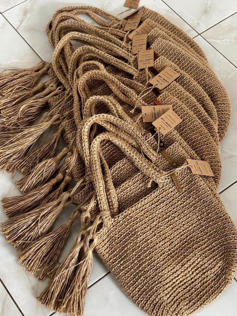 Puede incluir: Un conjunto de bolsas de mano de yute tejido. Cada bolsa tiene un asa trenzada y una borla decorativa. Las bolsas son de color beige natural y tienen peque&ntilde;as etiquetas adjuntas. Estas bolsas son adecuadas para llevar comestibles u otros art&iacute;culos.