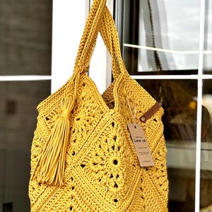 May include: Mustard yellow crochet tote bag with a geometric pattern and long braided handles. The bag features a large tassel and a small tag. The bag is hanging in front of a window.