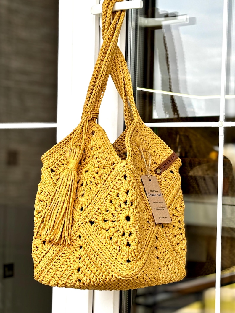 May include: Mustard yellow crochet tote bag with a geometric pattern and long braided handles. The bag features a large tassel and a small tag. The bag is hanging in front of a window.