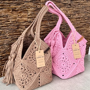 May include: Two handmade crochet bags, one in beige and one in pink, featuring a square motif pattern, sturdy handles, and a decorative tassel on the side. Each bag includes a brown paper tag with the text Lunar cat and a small leather label.