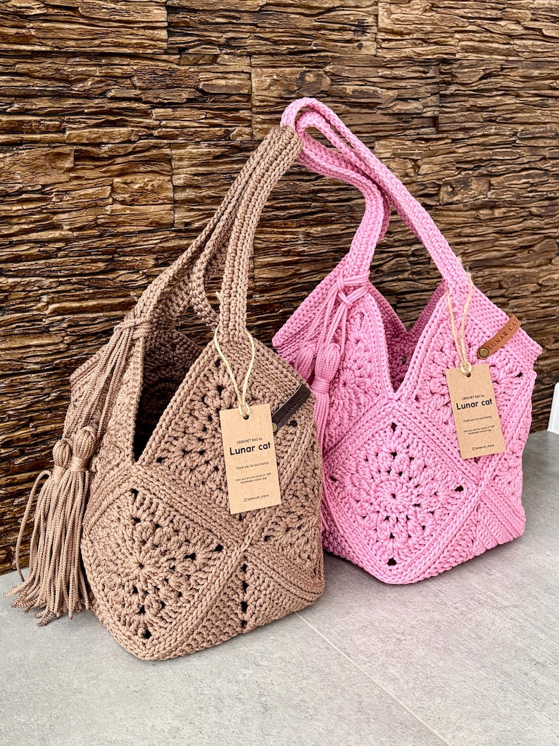 May include: Two handmade crochet bags, one in beige and one in pink, featuring a square motif pattern, sturdy handles, and a decorative tassel on the side. Each bag includes a brown paper tag with the text Lunar cat and a small leather label.
