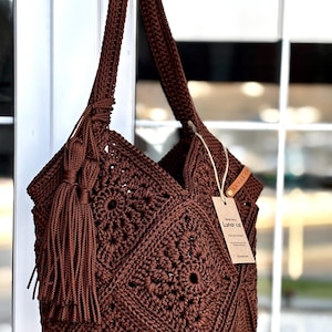 May include: A dark brown crochet shoulder bag featuring a square motif pattern, a long strap, and a large matching tassel attached to the side. A rectangular tan paper tag with the text Lunar cat is tied to the bag with a thin string.