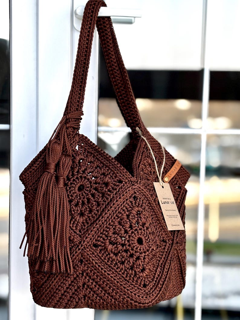 May include: A dark brown crochet shoulder bag featuring a square motif pattern, a long strap, and a large matching tassel attached to the side. A rectangular tan paper tag with the text Lunar cat is tied to the bag with a thin string.
