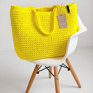 May include: A bright yellow crocheted tote bag with leather handles sits on a white chair with wooden legs. The bag has a small brown tag attached to it that says "Thank you for purchasing from The Cozy Corner. We hope you enjoy your new crocheted bag. Handmade with love."