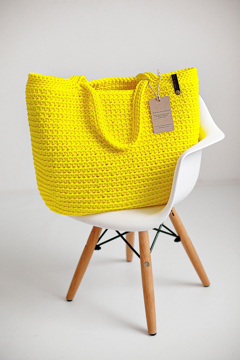 May include: A bright yellow crocheted tote bag with leather handles sits on a white chair with wooden legs. The bag has a small brown tag attached to it that says "Thank you for purchasing from The Cozy Corner. We hope you enjoy your new crocheted bag. Handmade with love."