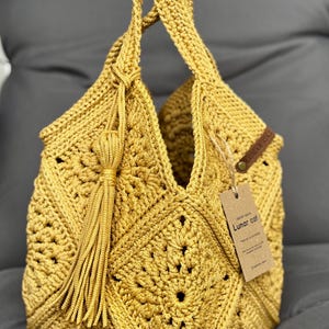 May include: Mustard yellow crochet bag with a geometric pattern and a tassel. The bag has two handles and a tag that reads "Lunar Cat". The bag is made of a soft, textured yarn.