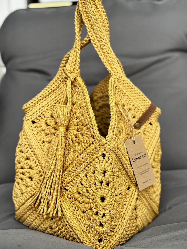 May include: Mustard yellow crochet bag with a geometric pattern and a tassel. The bag has two handles and a tag that reads "Lunar Cat". The bag is made of a soft, textured yarn.