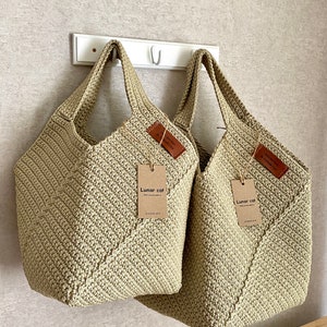 May include: Two beige crocheted tote bags hanging on a white hook. The bags have leather tags with the text "Lunar cat" and "Crochet bag by Lunar cat".