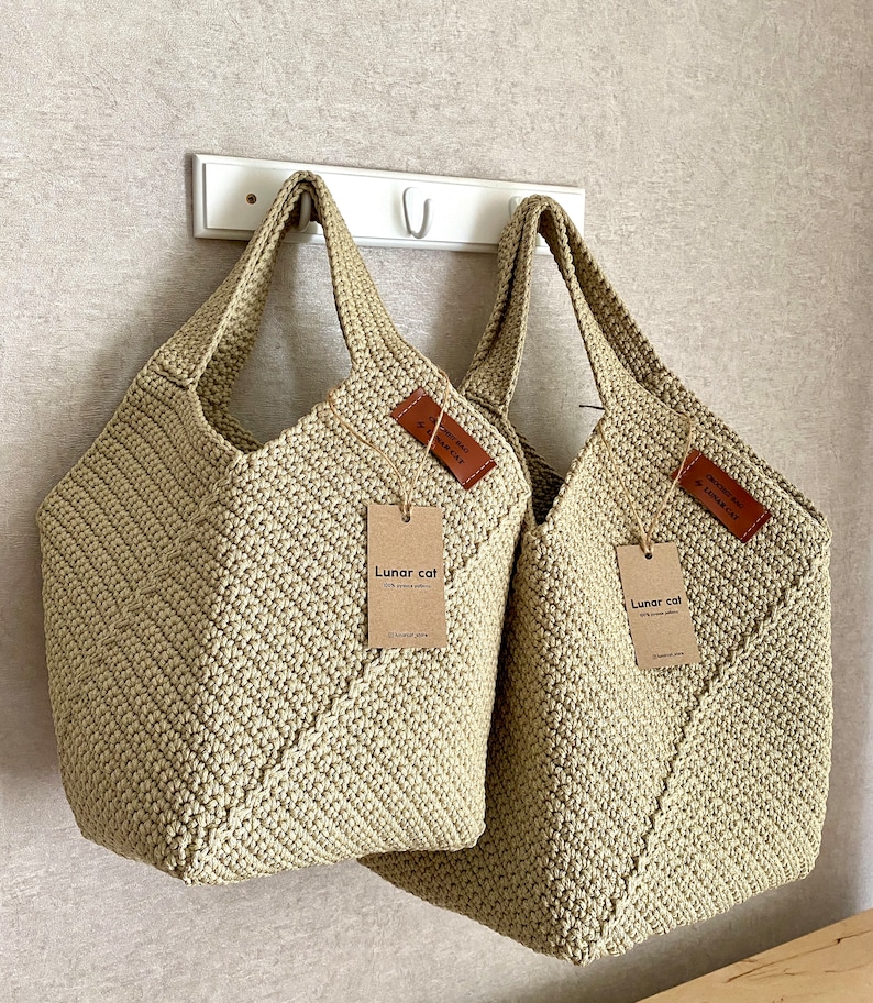 May include: Two beige crocheted tote bags hanging on a white hook. The bags have leather tags with the text "Lunar cat" and "Crochet bag by Lunar cat".
