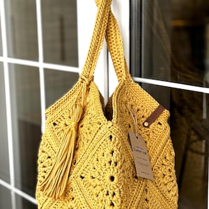 May include: Mustard yellow crochet tote bag with a diamond pattern and long handles. A long tassel hangs from the side. A tag with the text "Lunar cat" is attached to the bag. The bag is hanging in front of a white window.