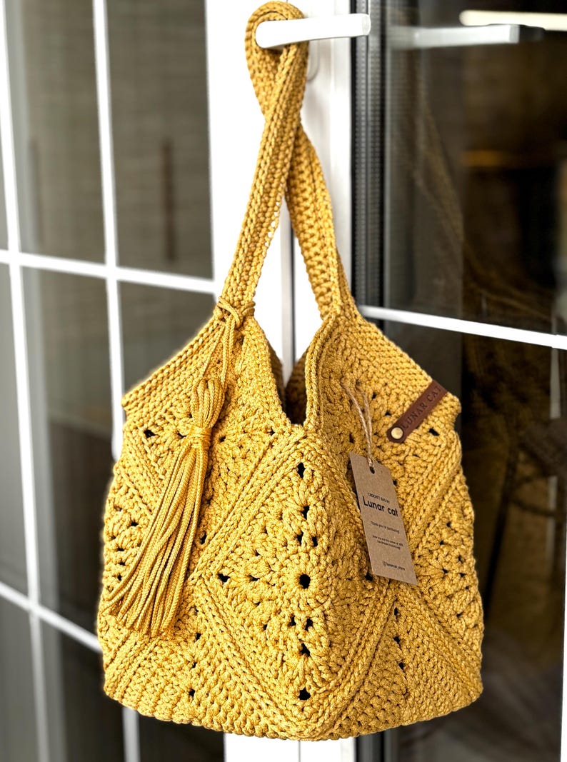 May include: Mustard yellow crochet tote bag with a diamond pattern and long handles. A long tassel hangs from the side. A tag with the text "Lunar cat" is attached to the bag. The bag is hanging in front of a white window.