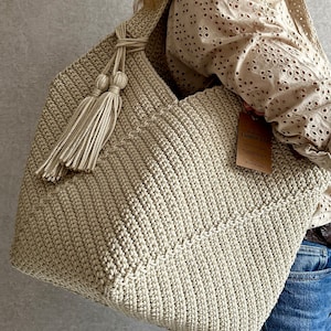 May include: A large, beige crocheted tote bag with a long tassel. The bag has a simple, textured design and is made with thick yarn.
