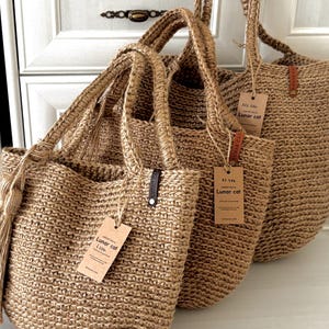 Puede incluir: Tres bolsas de mano de yute tejido de diferentes tama&ntilde;os. Las bolsas tienen asas resistentes y etiquetas adjuntas. Las bolsas son de color beige natural y parecen hechas a mano. El fondo es un armario blanco.