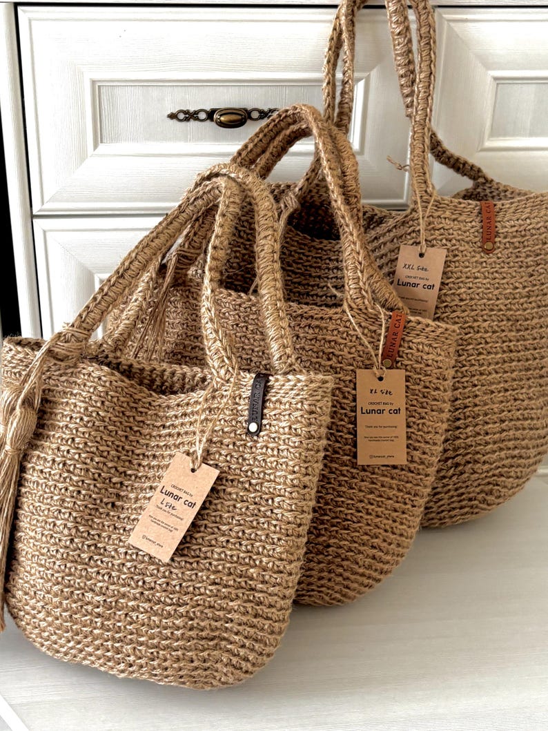 Puede incluir: Tres bolsas de mano de yute tejido de diferentes tama&ntilde;os. Las bolsas tienen asas resistentes y etiquetas adjuntas. Las bolsas son de color beige natural y parecen hechas a mano. El fondo es un armario blanco.