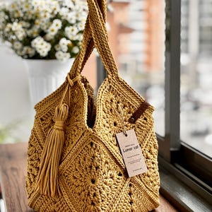 May include: Mustard yellow crochet tote bag with a geometric pattern and a long tassel. The bag has two handles and a tag that reads "Lunar cat". The bag is sitting on a wooden surface.