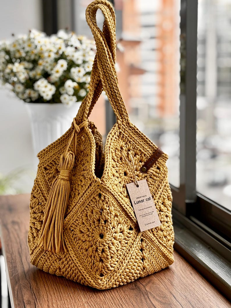 May include: Mustard yellow crochet tote bag with a geometric pattern and a long tassel. The bag has two handles and a tag that reads "Lunar cat". The bag is sitting on a wooden surface.