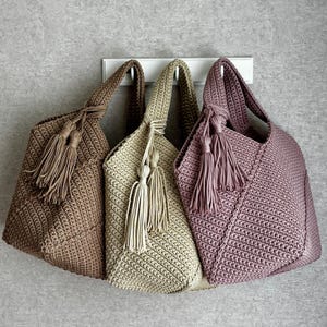 May include: Three crochet tote bags in shades of brown, beige, and mauve hang against a gray wall. Each bag features a knotted handle and decorative tassels. The bags are made of a textured, woven material.